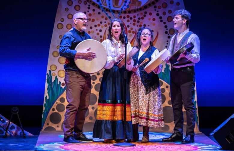 Four performers stand on a stage hold hand drums and singing into a microphone. Behind them, the stage is decorated in an intricate design of swirls and circles. 