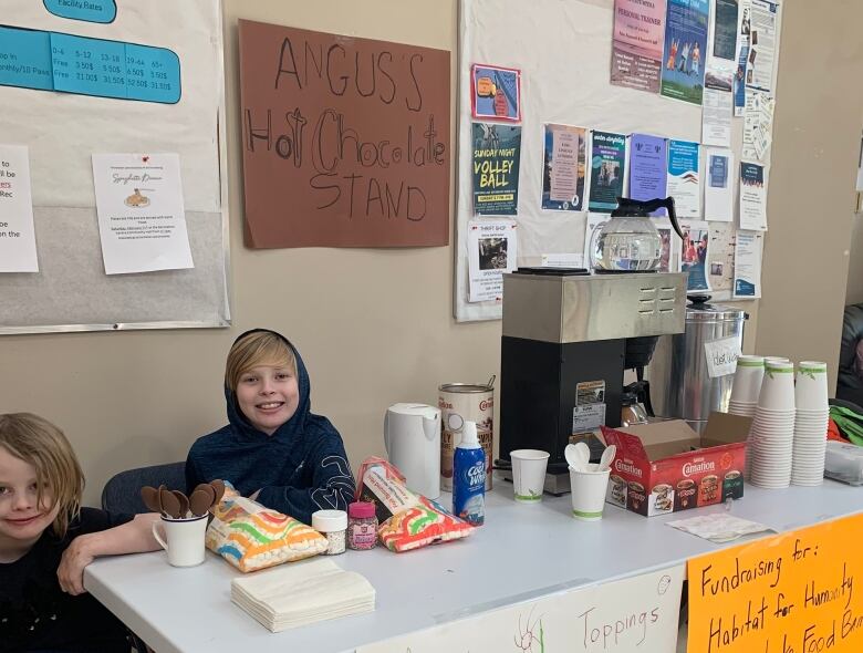 Two young boys sit at a table with signs taped to it, selling hot chocolate.