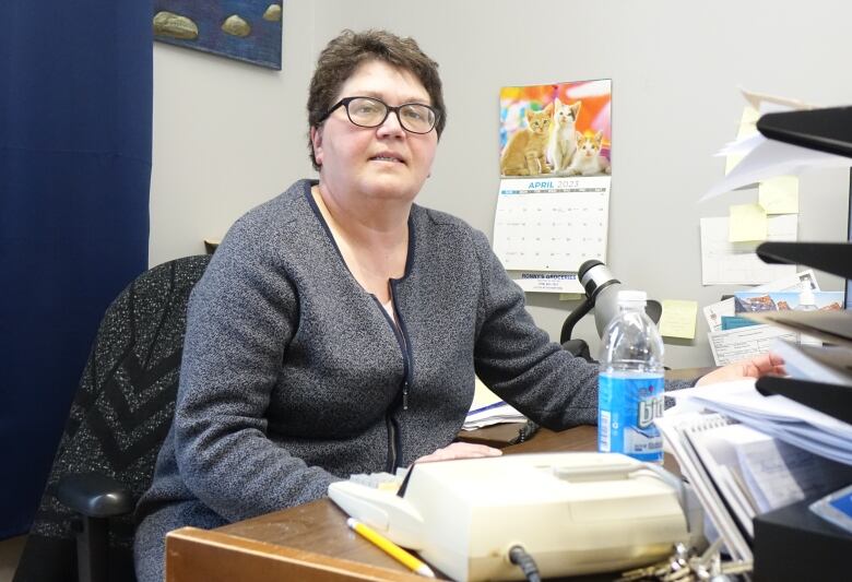 A woman in glasses and a grey sweater sits at a cluttered desk.