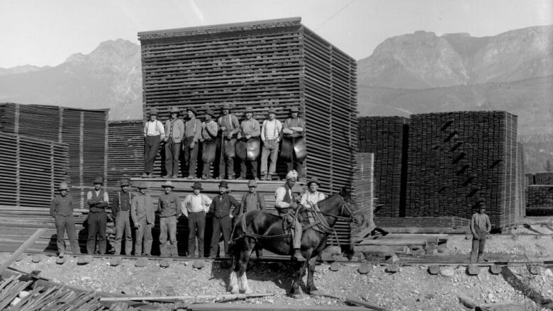 A black-and-white photo where a man is riding on a horse in front of a group of men standing amid piles of lumber, with hills in the background.