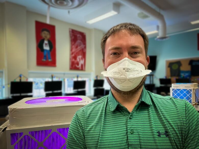 A man with short dark hair wearing an N95 mask and green collared shit with navy stripes standing in front of two homemade air purifiers systems, one purple, the other blue.