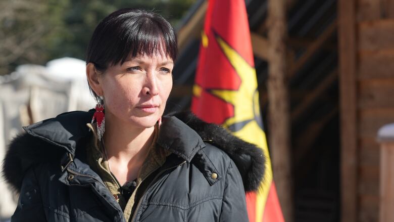 A woman with dark hair, wearing a black winter jacket and beaded earrings, is shown outside a small shed adorned with a red, yellow and black flag.