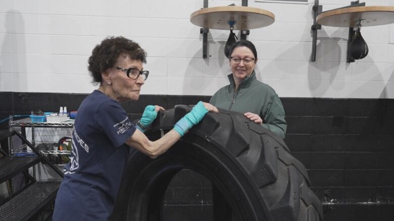 Two woman stand around a large tire.