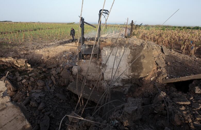 A man stands in a field beside a crater, caused by an airstrike. The crater reveals concrete rubble and twisted metal.