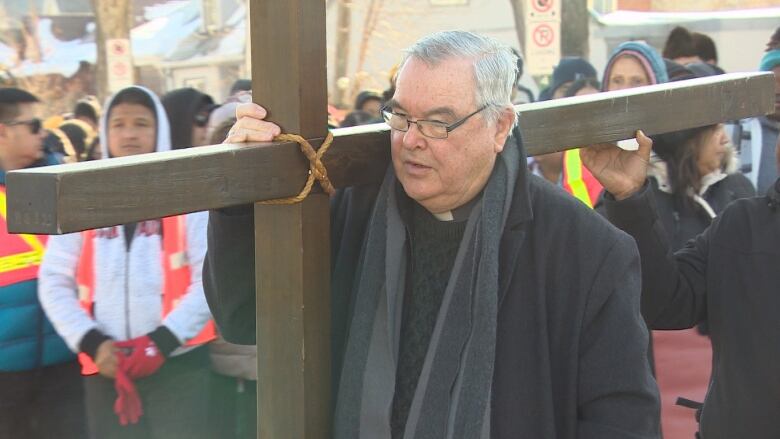 A man in short white hair and glasses, and wearing a long black coat, carries a wooden cross on his right shoulder as a crowd walks behind.