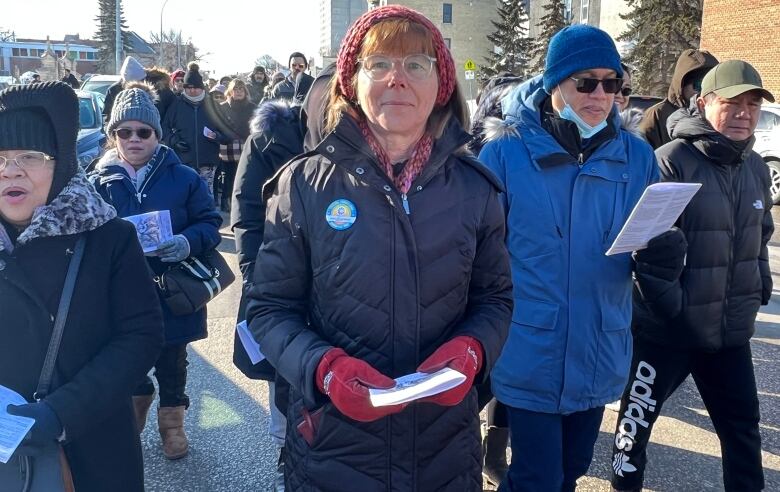 People walk, holding sheets of paper and singing. One woman, wearing a knitted headband, long coat and eyeglasses, smiles at the camera.