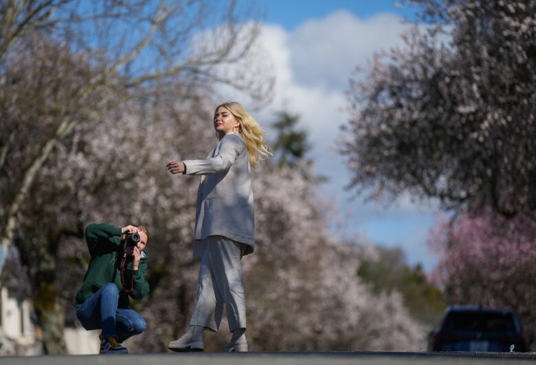 A blonde woman poses for a picture in the middle of a street with cherry blossoms on it, with a man crouching behind her to take a picture with a camera.