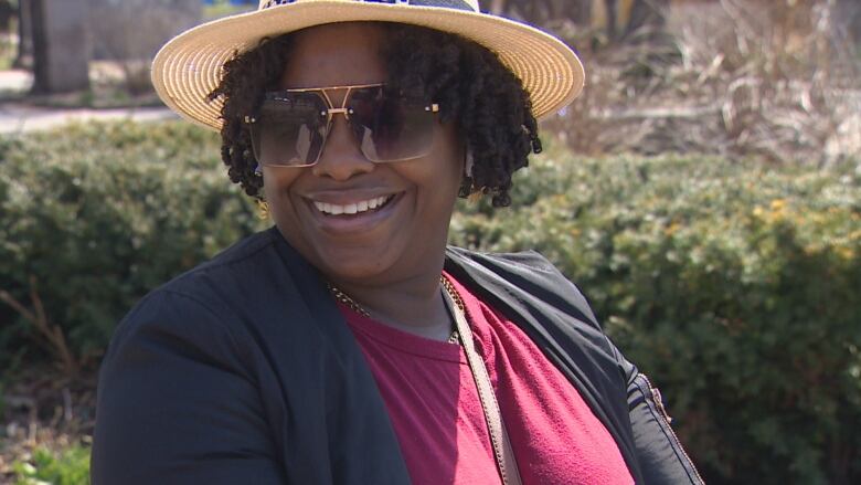 A woman in a tan hat, sunglasses and pink shirt smiles at the camera. 