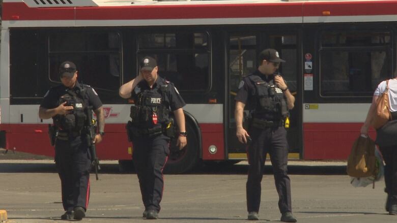 Three police officers stand in the foreground. A TTC bus is in the background. 