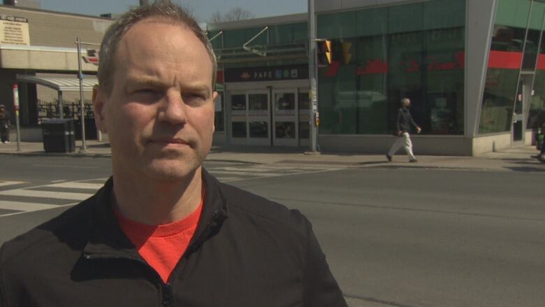 A man in a light black jacket and red shirt stands in front of an intersection with a TTC station in the background. 