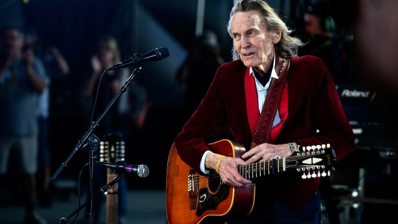 An older man wearing a red blazer and standing in front of a microphone holds a 12-string guitar in front of an audience. 