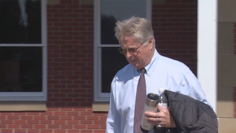 A man in a shirt and tie walking with a jacket, a plastic water bottle and a thermos in his arms