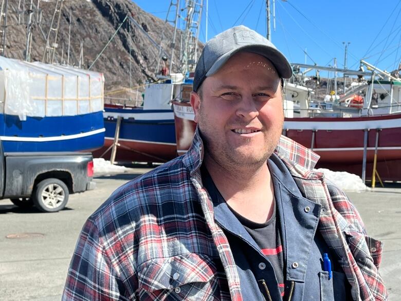 A man in a baseball cap and plaid jacket stands in front of small fishing boats.