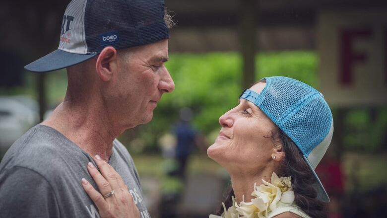 A man and a woman in backward-turned baseball caps smile while facing each other, she with her hands on his chest.