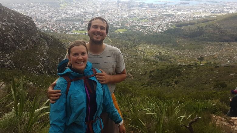 A man with brown hair and grey shirt holds the shoulders of a woman with light brown hair and a blue jacket on top of a mountain. 