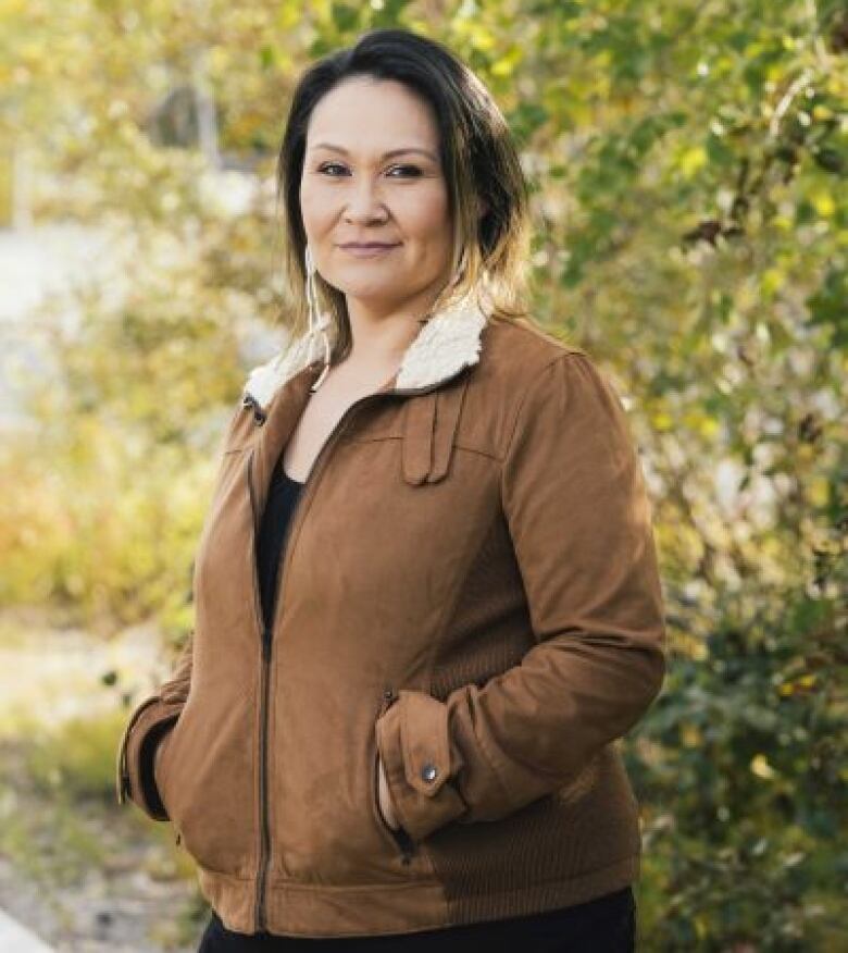 A woman stands in the outdoors wearing a brown jacket. 