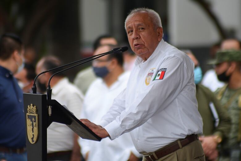 A man in a white shirt is shown speaking into a microphone at a podium.