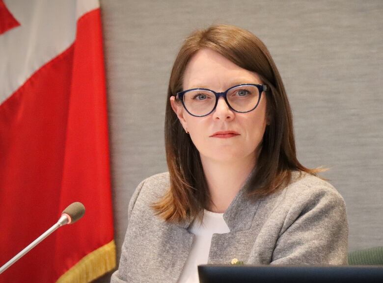 A woman with glasses and a grey jacket looks off with a Canadian flag on the wall behind her.