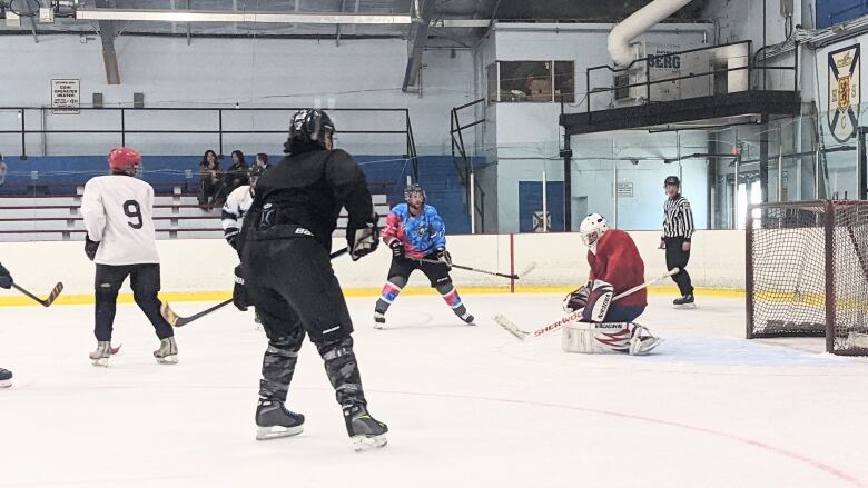 Four hockey players are facing a goaltender, who is down on the ice in the butterfly position in front of their net. A referee stands behind the net.