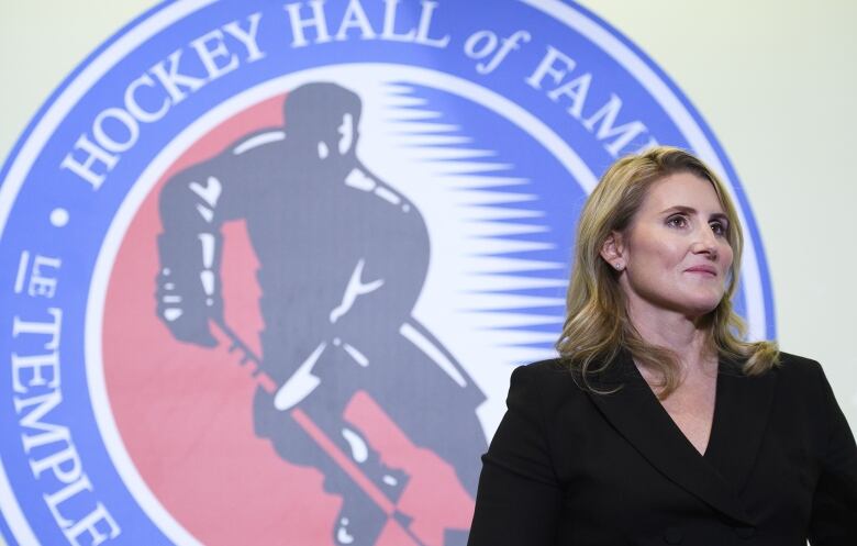 A women's hockey player stands in front of a Hockey Hall of Fame logo.