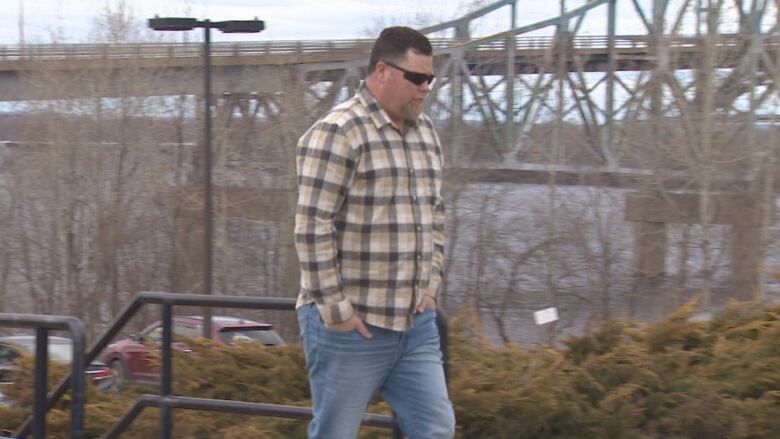 A man wearing a plaid shirt and sunglasses walks with a bridge in the background.