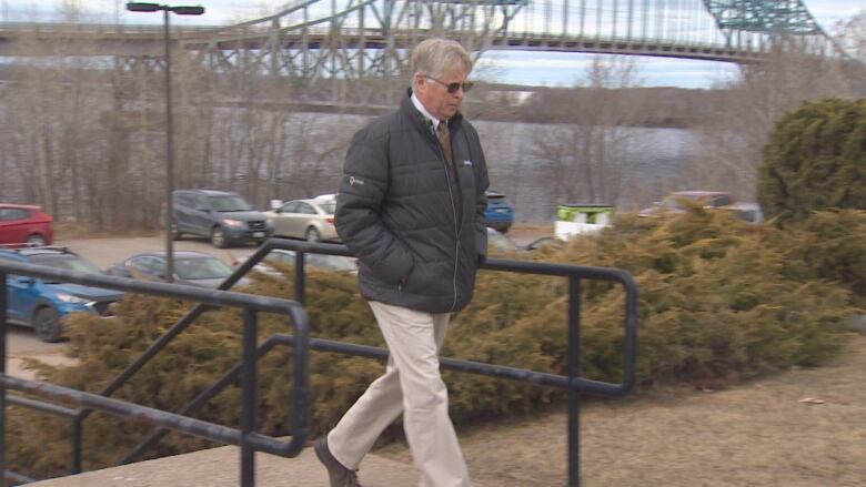 A man wearing a black jacket and sunglasses walks with a bridge in the background.