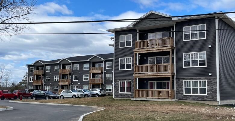 A grey apartment complex with wooden balconies and parking lot filled with cars on the left.