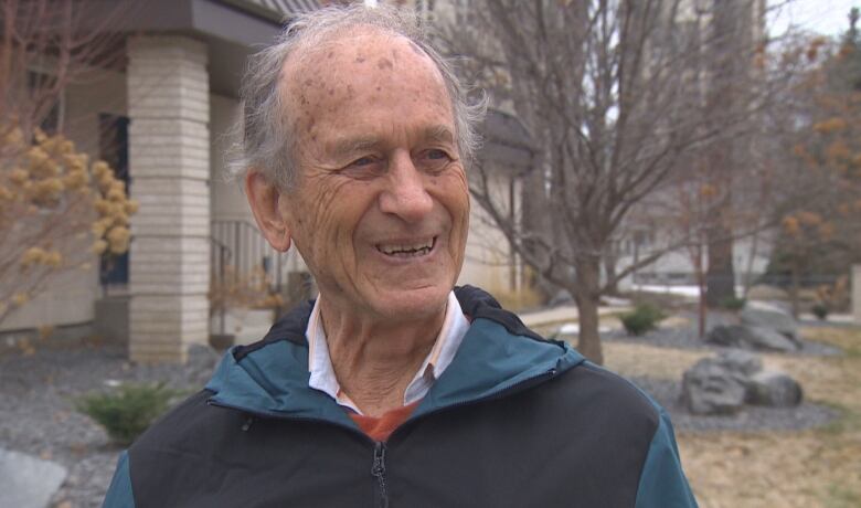 An older man, with thinning white hair, almost bald, stands outside, smiling and looking to the right of the photo. It's a cloudy day and behind him are trees without their spring leaves and grass just revealed from snow melt.