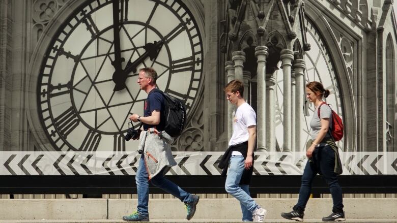 Three people walk in front of a large black-and-white image of an old clock on a tower.