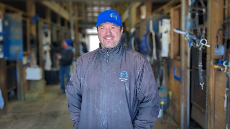 A man in a blue hat stands in a barn full of horses 