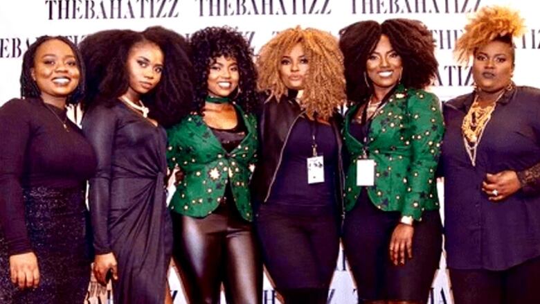 Six brown-skinned women stand smiling in front of a white backdrop which says THEBAHATIZZ in black.