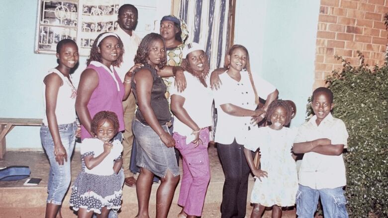 Eight children stand in a line in front of a house, smiling at the camera. A man and woman stand behind them, smiling.