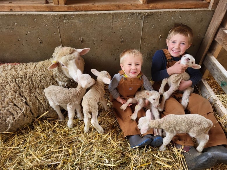 Two children surrounded by lambs smile while the mother lamb looks on.