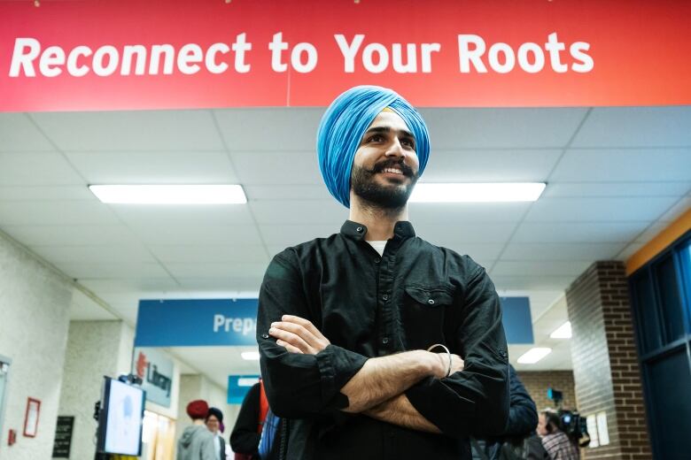 Man wearing black button up shirt and blue turban stands in front of a sign that reads 