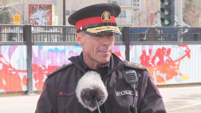 The police chief wears a black and red police hat and speaks before a large, fluffy microphone. Behind him is a colourful floral mural. 