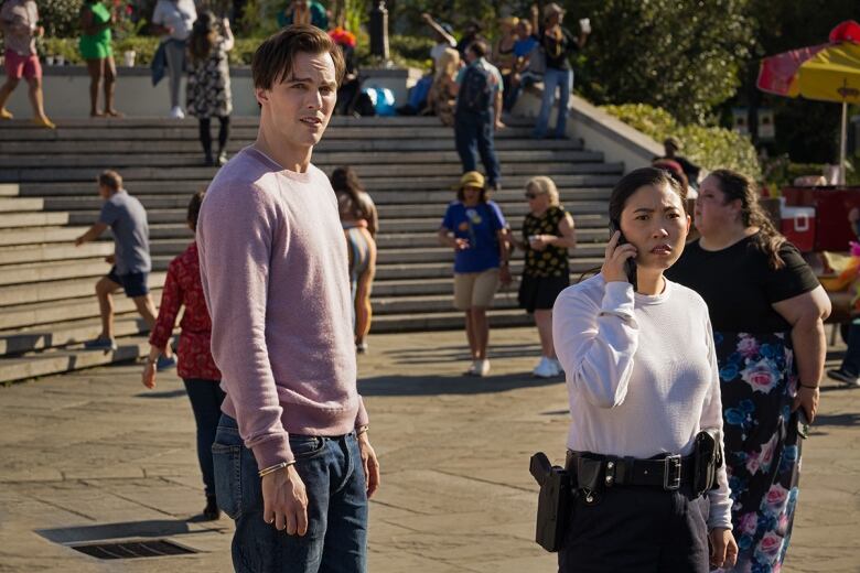 A man and a woman stand in a small amphitheatre. The man is wearing handcuffs, though his arms hang freely as the handcuffs are not attached to one another. The woman has a utility belt with police equipment. The woman is speaking on a cellphone. Both are looking quizzically into the distance.