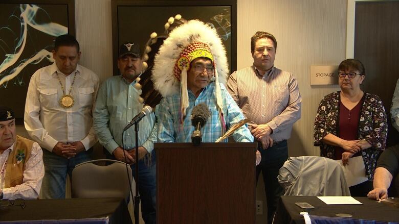 A chief wearing a headdress speaks at at a podium. 