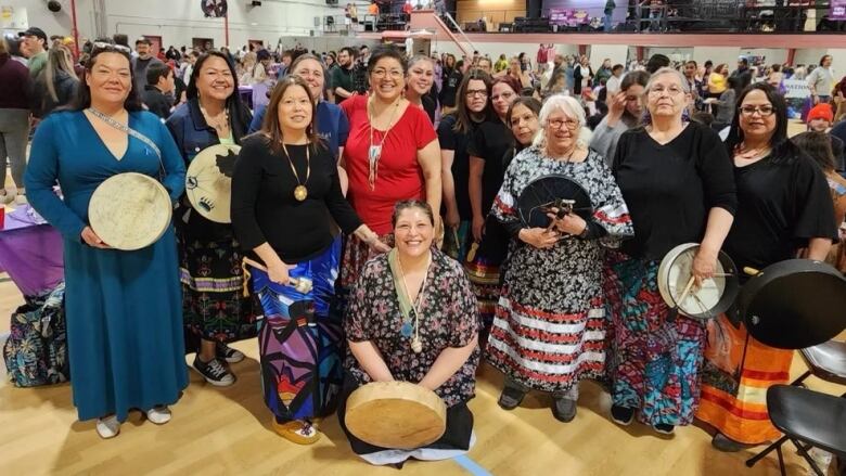 A group of women wearing colourful dresses and skirts stand together holding hand drums.