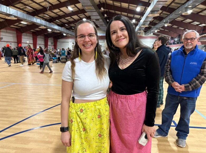 A woman wearing a white t-shirt and yellow skirt and a woman wearing a black shirt and pink skirt stand together smiling.