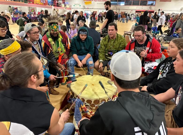 A group of people sit in a circle around a drum, each holding a drumstick.