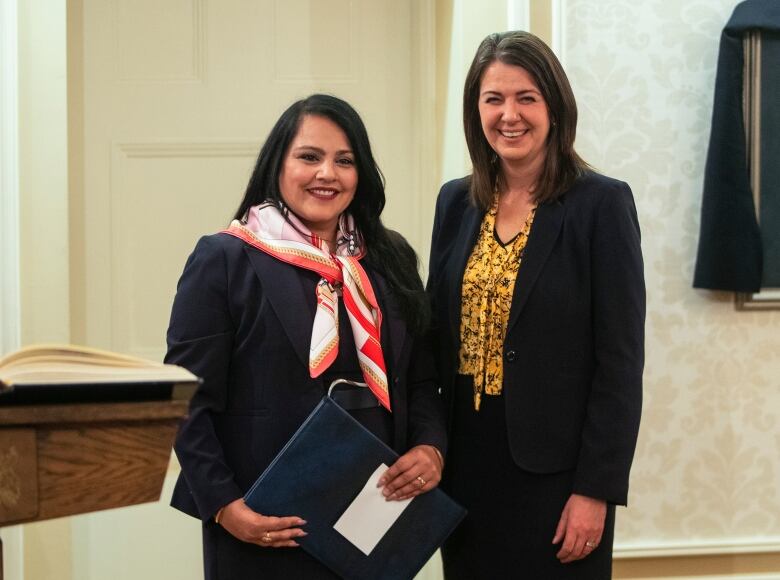 Newly appointed cabinet minister Rajan Sawhney holds a folder while standing next to Alberta Premier Danielle Smith at the cabinet swearing-in ceremony. Both women are smiling.