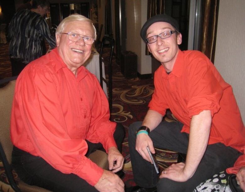 Two men sit facing each other looking at the camera. They're wearing red matching dress shirts.