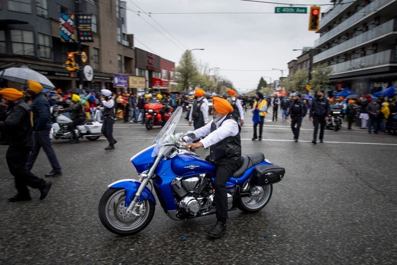 A Sikh man in a motorcycle leads a parade of people behind him on a rainy street.