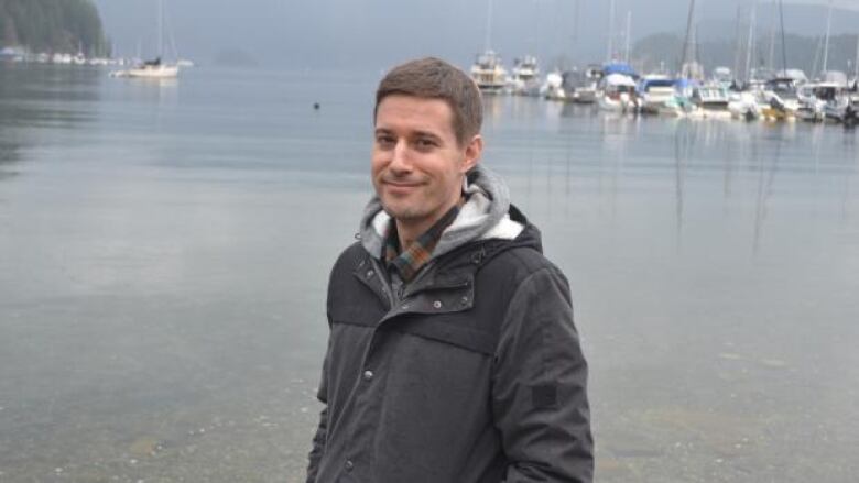 A man with short hair wearing a dark grey parka standing in front of a marina and smiling at the camera