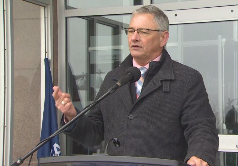 A man in glasses and dark jacket stands behind a microphone in front of glass doors.