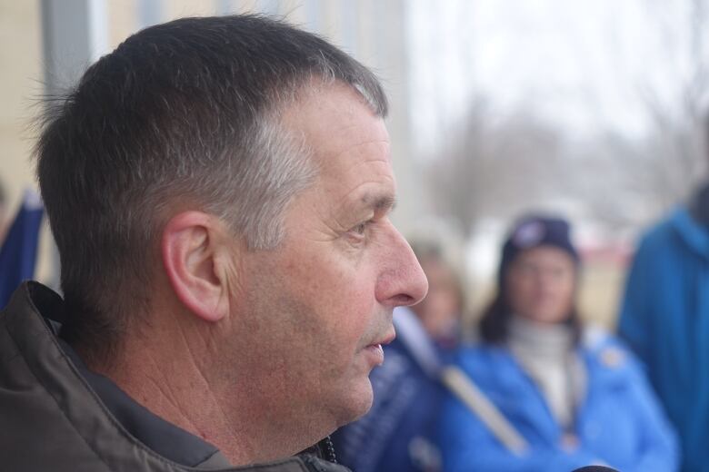 A headshot of a man speaking, with people watching in the background.