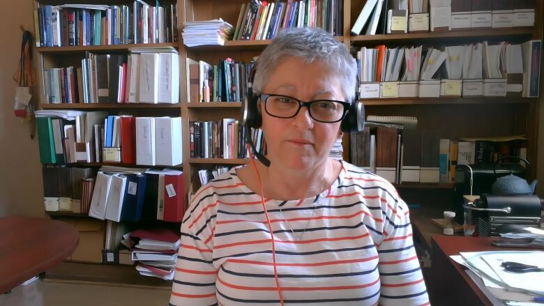 A woman is shown wearing headphones on a video call with a bookcase behind her.