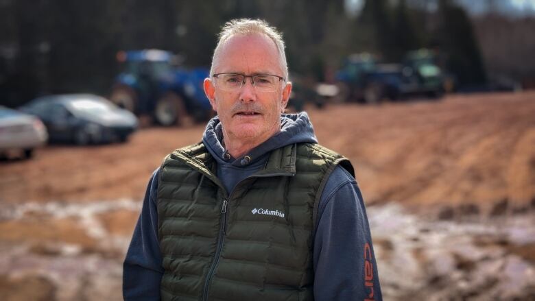 A man wearing a hoodie and green vest stands in a farm yard on P.E.I. 