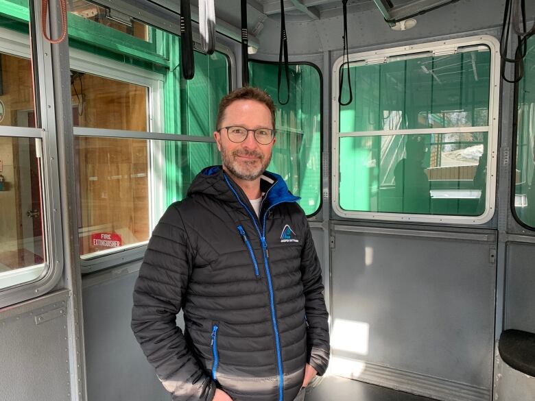 A man with glasses and waring a black jacket stands in a cablecar.