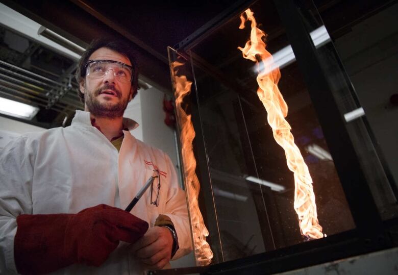 A man with dark hair and a beard wears protective equipment as he stands next to a vertical column of flame behind a glass case.